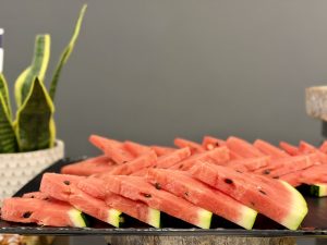 A platter of neatly arranged watermelon slices sits on a black tray. The slices are cut into triangular shapes, revealing their bright pink, juicy flesh dotted with small black seeds. The outer green rind is still visible at the base of each slice. In the background, there is a green potted plant with upright leaves, adding a touch of color and freshness to the scene, which is set against a simple gray backdrop.