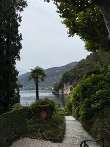 Curving stone path through green trees and bushes leading to a lake, with mountains in the background under a cloudy sky.