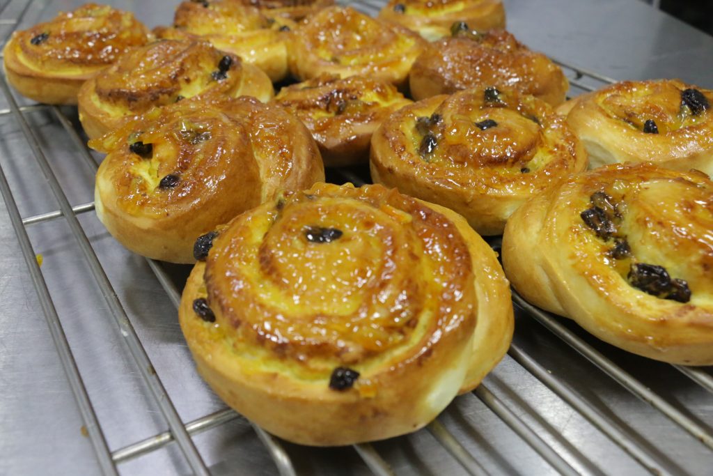 Golden-brown spiral cinnamon roll buns with raisins and a glossy glaze cooling on a metal wire rack. Multiple freshly baked buns are arranged closely together.