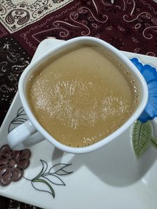 A creamy Tea in a white cup on a decorative plate with floral patterns, placed on a richly patterned maroon tablecloth. 
