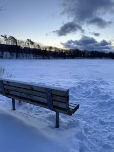 A close-up, angled shot of a wooden bench covered in light frost, surrounded by thick snow footprints. In the background, a vast frozen field or lake stretches toward a distant tree line under a blue and gray sky with dark, heavy clouds at dusk. 
