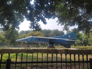 A vintage Indian Air Force fighter jet on static display in a tree lined park behind an iron fence.
