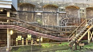 A dense bundle of cables and conduits runs along industrial metal trays beside a weathered brick viaduct. Pink and black cables curve toward a circular junction on the right, flanked by a metal staircase. Dim lights and patches of moss add to the gritty, urban atmosphere.