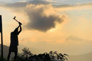 A dark silhouette of a person raises an axe or hammer above their head, appearing ready to strike, set against a dramatic sky glowing with golden light from the sun behind clouds. Hills and vegetation form the backdrop. 