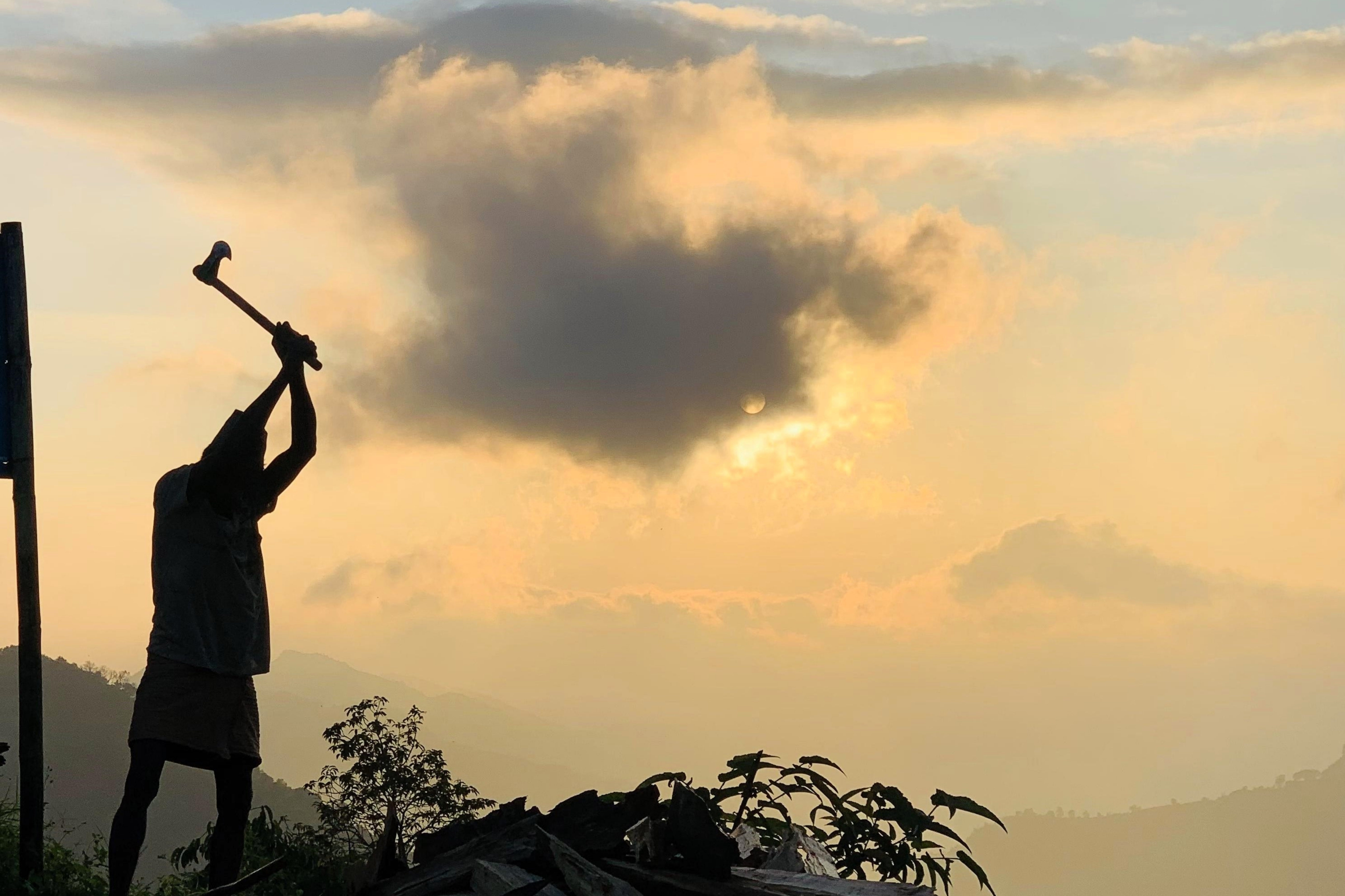A dark silhouette of a person raises an axe or hammer above their head, appearing ready to strike, set against a dramatic sky glowing with golden light from the sun behind clouds. Hills and vegetation form the backdrop.