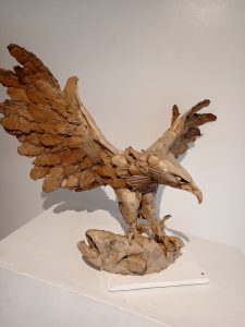 A bird sculpture with spread wings standing on a fish on a white display stand.
