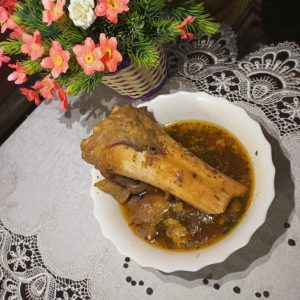 A plate with a large, cooked bone piece in savory brown gravy, set on a lace tablecloth with a vibrant flower arrangement nearby.