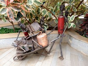 Metallic sculpture of figures working and pulling with a wheelbarrow filled with gas canisters, welded entirely from scrap metal parts like pipes and old tanks. 
