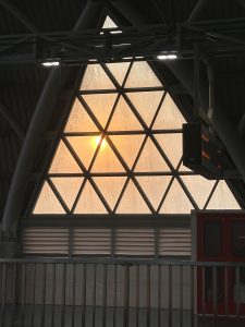 Triangular windows of Dhaka metro station with a geometric grid frame a soft orange sunset, casting a warm glow inside an industrial interior. A digital display hangs nearby. 
