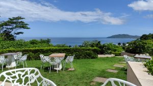 Outdoor seating area by the ocean with white chairs and tables on grass. Sunny sky, green surroundings, and a distant island view.