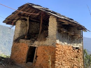 A rustic two‑story house made of stone and mud bricks with a wooden roof covered in flat stone slabs. Bundles of straw are stored under the eaves, and forested hills rise in the background beneath a clear blue sky.