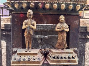 Two small gilded brass devotee figures in namaste prayer posture — a man in traditional Nepali daura suruwal and a woman in sari — mounted on a temple pillar base, Kathmandu, Nepal. 