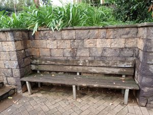 A long, rustic wooden bench with a multi-slat backrest and seat with vegetation and a garden behind it on an elevated platform above. 