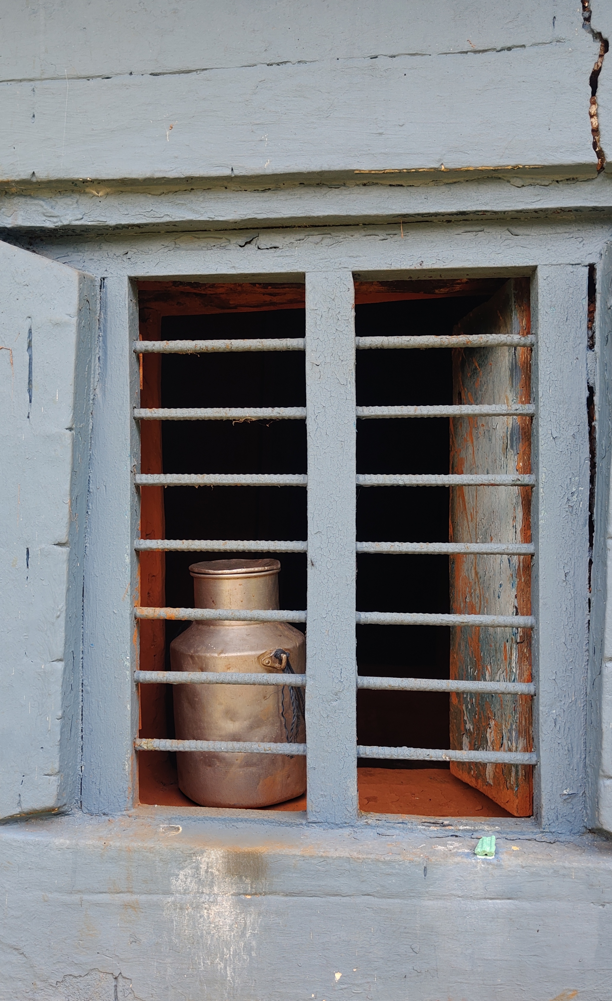 A weathered, metal milk can sits behind blue-gray bars of a rustic window.