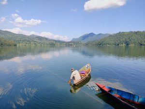 Fisherman pulling a net from a wooden boat on the glassy waters of Begnas Lake with forested hills beyond 