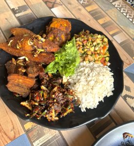 A mound of white beaten rice rests beside a dark, spiced mixture of marinated meat chunks tossed with green chilies and herbs. Crispy, seasoned potato wedges garnished with scallions sit near thick-cut pan-fried meat pieces, completed by a colorful peanut and soybean salad arranged around a single crisp green lettuce leaf.