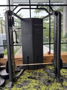 A dual-pulley cable crossover machine in a sunlit gym with a barbell and weights on a green floor.