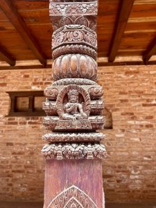 Close-up of an intricately carved wooden temple column featuring a meditating deity within a shrine niche, lotus motifs, and scrollwork, set against red brick. 