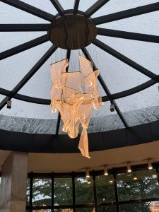 A chandelier hanging from the ceiling of a restaurant, with large windows and a decorative circular glass feature above.