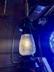 A vintage-style black metal kerosene lantern with glowing filament bulb and etched glass globe, hanging from a braided rope in a dimly lit space with blue ambient light. 