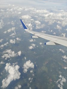 This aerial window shot features an airplane wing with distinct blue tips cutting through the upper right side of the frame. Below, a patchwork of green land is partially covered by a scattered blanket of fluffy white clouds.