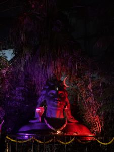 Illuminated statue of Lord Shiva with a large crescent moon, draped in glowing red and purple hues, surrounded by lush foliage in the night.