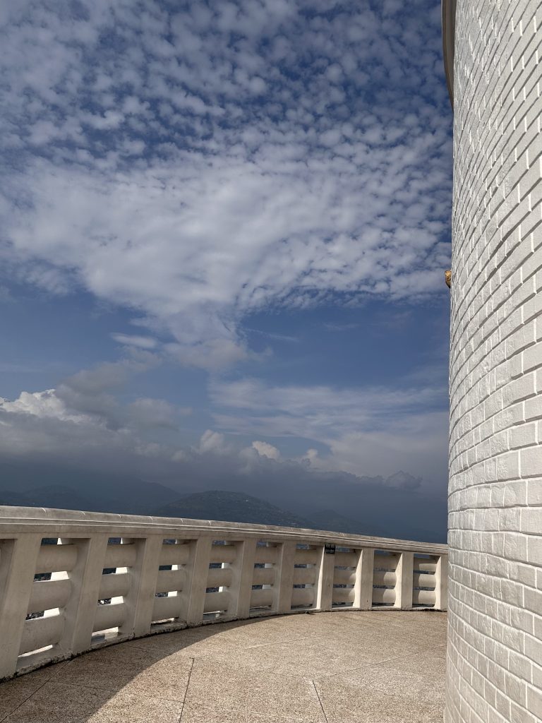 Curved white brick wall of Peace Stupa, Nepal and stone terrace with railing overlooking distant mountains under a partly cloudy sky.