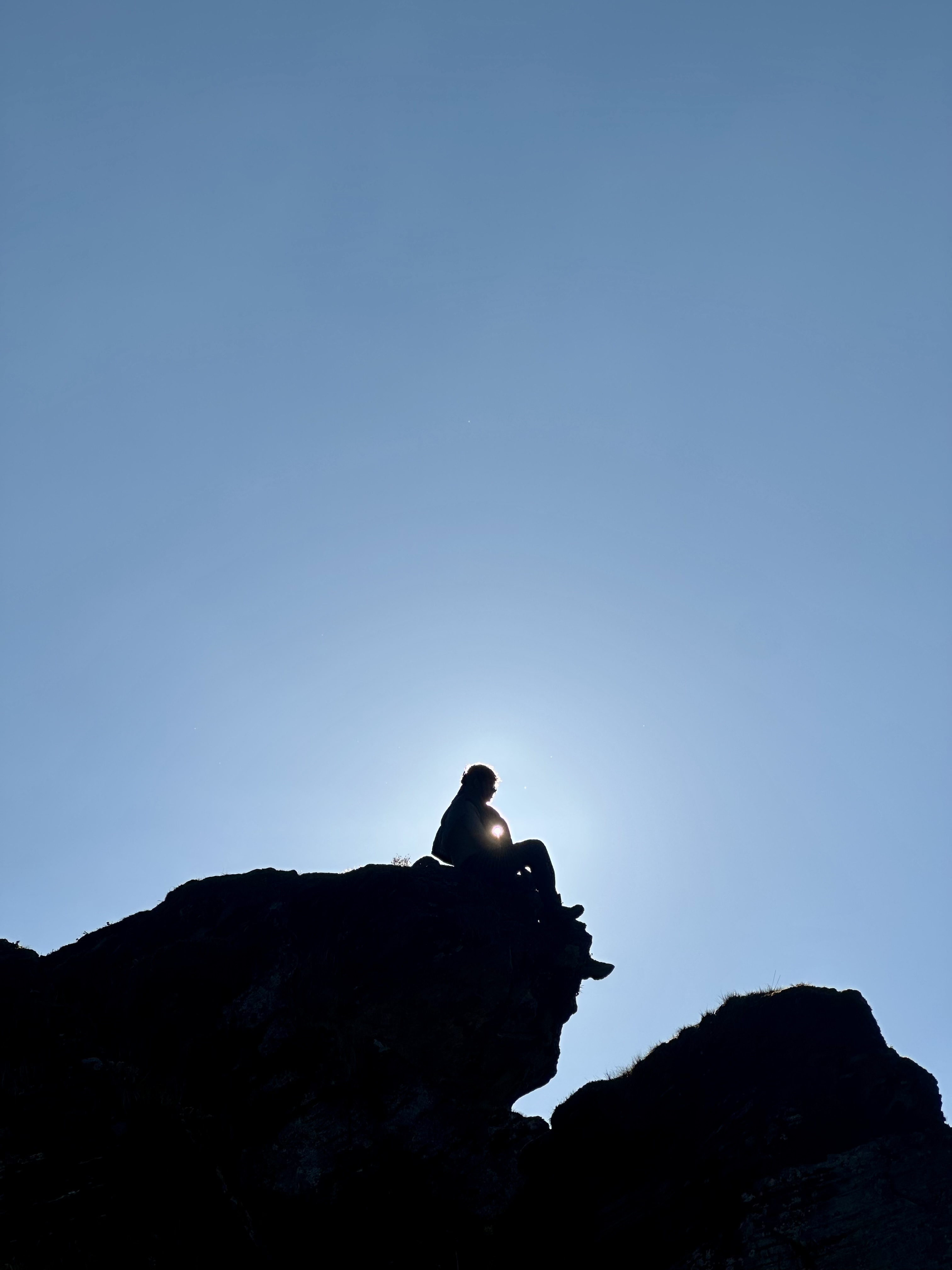 A solitary figure is silhouetted against a vast clear blue sky, seated atop a rugged rocky outcrop with the sun glowing directly behind them, creating a striking backlit halo effect.