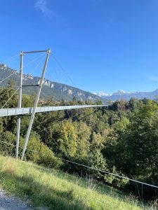 A long suspension bridge stretches over a lush green valley, with tall mountains and a clear blue sky in the background