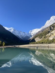 Snow-capped mountains reflect in a calm turquoise alpine lake in Manang. Surrounded by rocky cliffs and lush pine trees under a clear blue sky, the scene is peaceful, breathtaking, and majestic.