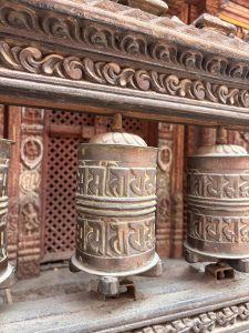 Close-up of aged brass Buddhist prayer wheels (mani wheels) engraved with Sanskrit mantras lined up beneath an ornately carved wooden temple beam, Nepal.