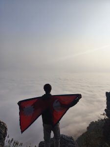 A person stands on a rocky cliff, holding the Nepal flag, overlooking a sea of clouds at sunrise. 