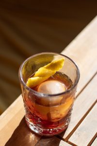 Close-up of an amber cocktail in a patterned glass on a sunlit wooden table, served with a large round ice cube and a twisted lemon peel garnish.
