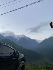 A dark pickup truck parked on a hillside overlooking a deep green valley, with a row of snow-capped Himalayan peaks visible in the distance under a partly cloudy sky. Power lines cross the upper portion of the frame, and a hand holding a phone is visible at the right edge.
