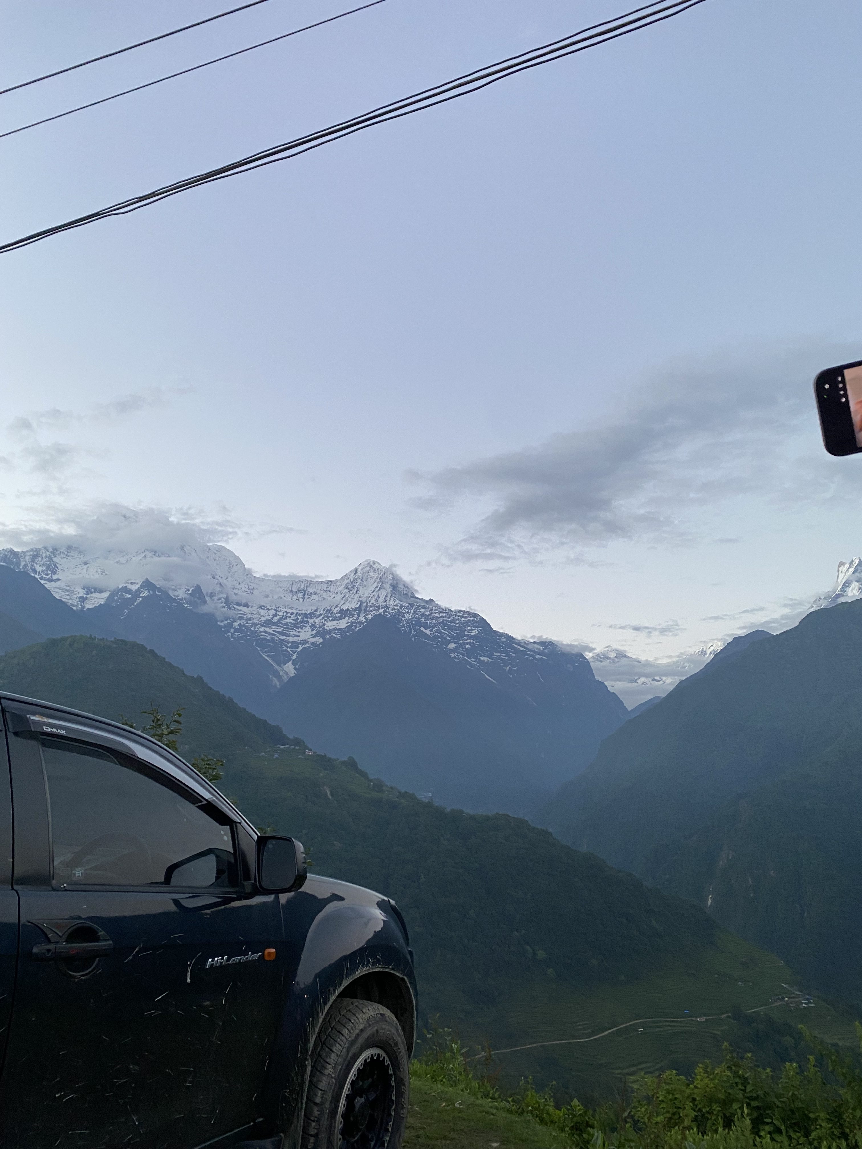 A dark pickup truck parked on a hillside overlooking a deep green valley, with a row of snow-capped Himalayan peaks visible in the distance under a partly cloudy sky. Power lines cross the upper portion of the frame, and a hand holding a phone is visible at the right edge.