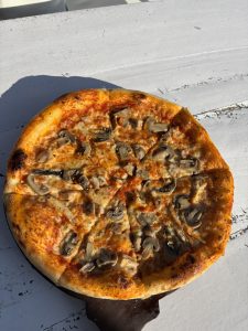 Wood-fired mushroom pizza with golden charred crust served on a rustic wooden board in bright afternoon sunlight 