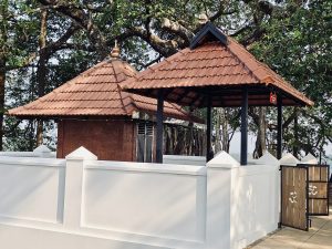 A traditional Kerala style shrine stands peacefully beneath a large banyan tree inside the temple grounds. The tiled roof and white compound wall reflect Kerala's timeless temple architecture. Captured at Niramkaitha Kotta Bhagavathi Temple, Vallikunnu North, Malappuram.
