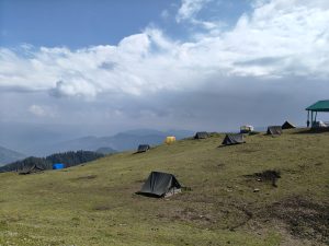 Camping tents on a grassy mountain ridge in India under dramatic clouds with panoramic Himalayan views
