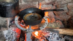 A blackened frying pan heats over an open flame on a rustic stone hearth.
