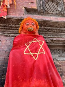 A brightly painted red deity statue, possibly Hanuman, covered in vermillion and adorned with marigold garlands and a red cloth bearing a gold geometric star symbol, Patan Durbar Square, Nepal. 