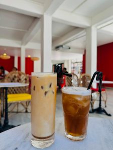 A tall glass of iced milk coffee and a refreshing glass of iced tea, both served with curly black straws, sitting on a white table inside a bright café featuring bold red walls and woven wicker chairs. 