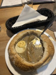 A traditional bread bowl filled with hearty soup, topped with a boiled egg and chunks of meat. Served on a wooden table, it offers a warm and rustic dining experience.
