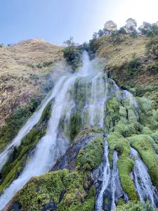 A low-angle shot of a tall, powerful waterfall flowing down a rugged mountain slope. The water divides into multiple streams as it cascades over dark rocks heavily blanketed with bright green moss and vegetation. The top of the hill is lined with sparse trees under a clear sky. 