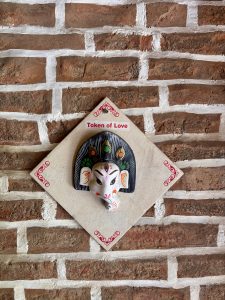 A hand-painted Ganesha face mask mounted on a diamond-shaped lokta paper board labeled “Token of Love,” hung on an exposed brick wall.