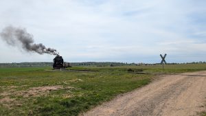 The Brockenbahn steam train travels through a wide green landscape, leaving a trail of dark smoke as it approaches a rural railway crossing.