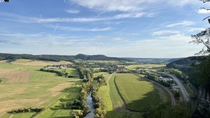 A sweeping rural landscape of rolling hills, patchwork fields, and a winding tree-lined river under a clear blue sky. A cluster of buildings and a curving road in the foreground hint at a nearby village amid the peaceful countryside.
