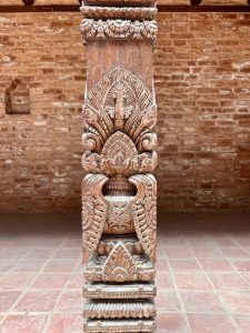 A richly carved Newari temple wooden pillar featuring a fierce face (likely Kirtimukha), lotus petals, and elaborate foliate scroll carvings, standing on a tiled courtyard floor against a brick wall. 