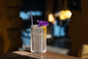 Iced cocktail in a textured glass on a wooden table, garnished with purple flowers and a lemon slice, with a dark blurred bar interior in the background.
