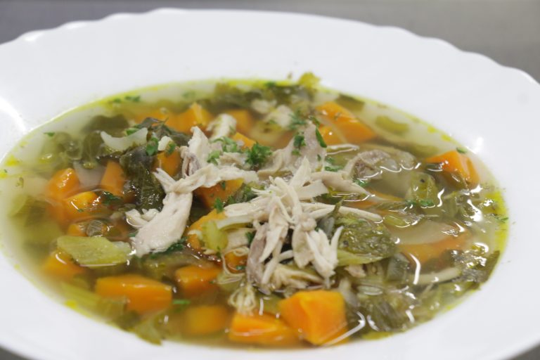 A bowl of chicken soup with carrots, greens, and herbs in a clear broth, served on a white plate.