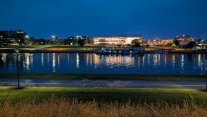 Nighttime view across a river with city lights reflecting on the water, a boat docked near the shore, and a brightly lit modern building in the background.